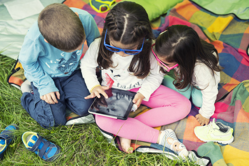 Three children with colorful sunglasses playing with a digital tablet on a picnic blanket under a camping tent.