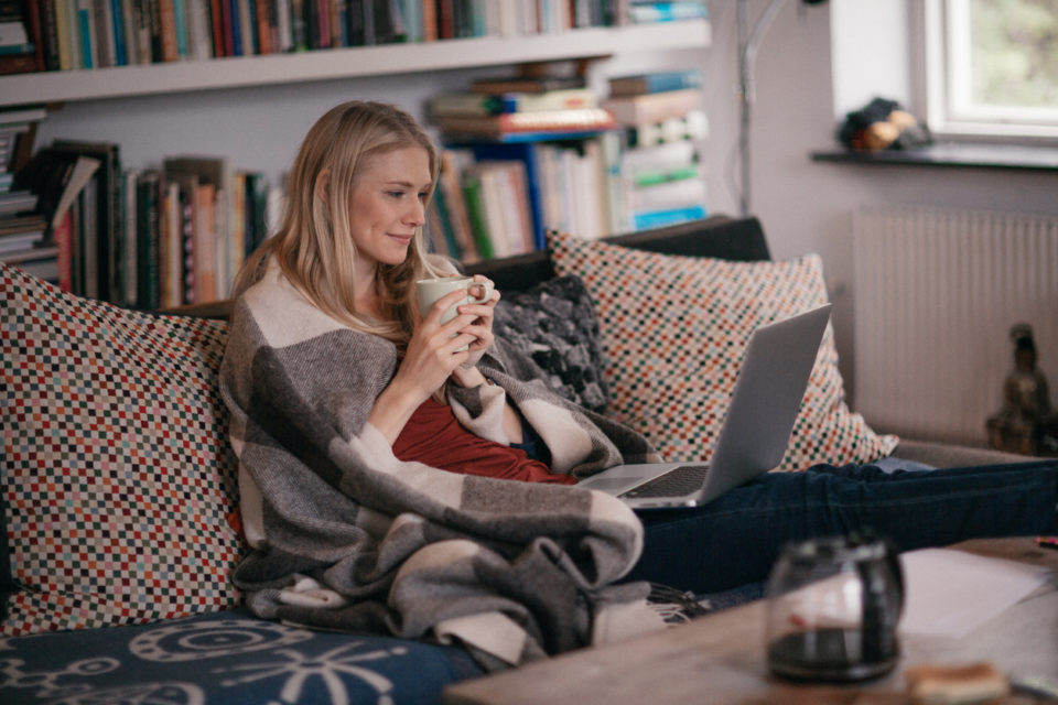 Woman sitting on her couch wrapped in a blanket with a laptop open in front of her