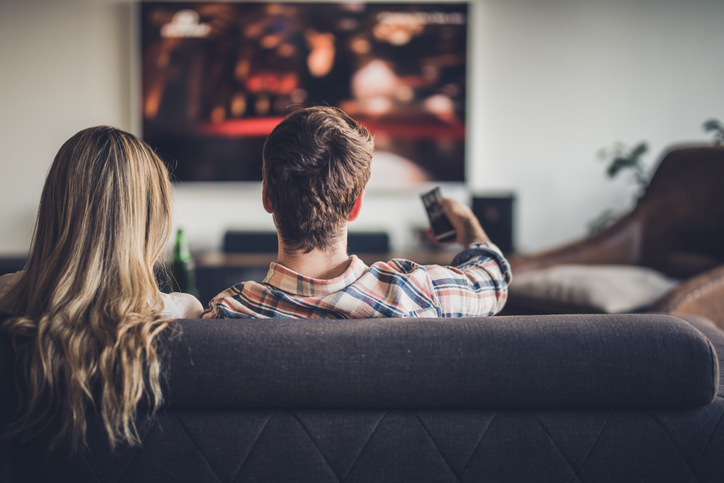 Back view of a couple watching TV on sofa in the living room. Man is changing channels.