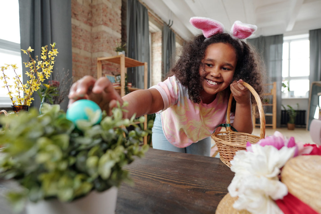 Happy little girl with toothy smile putting painted Easter egg into flowerpot